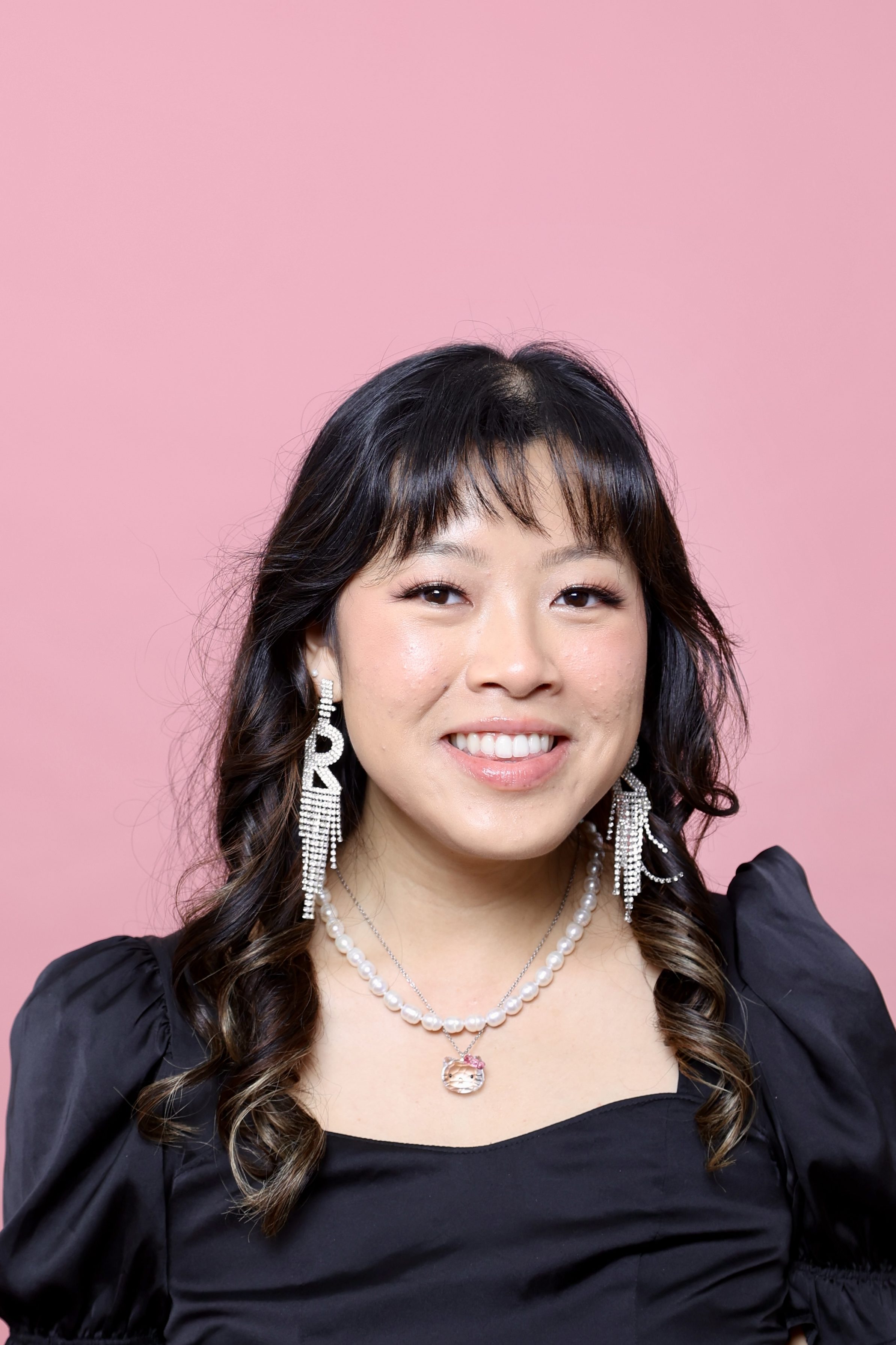 headshot of robin with a pink background, she's wearing a black sweetheart neckline short puff sleeved shirt and silver crystal "R" earrings.