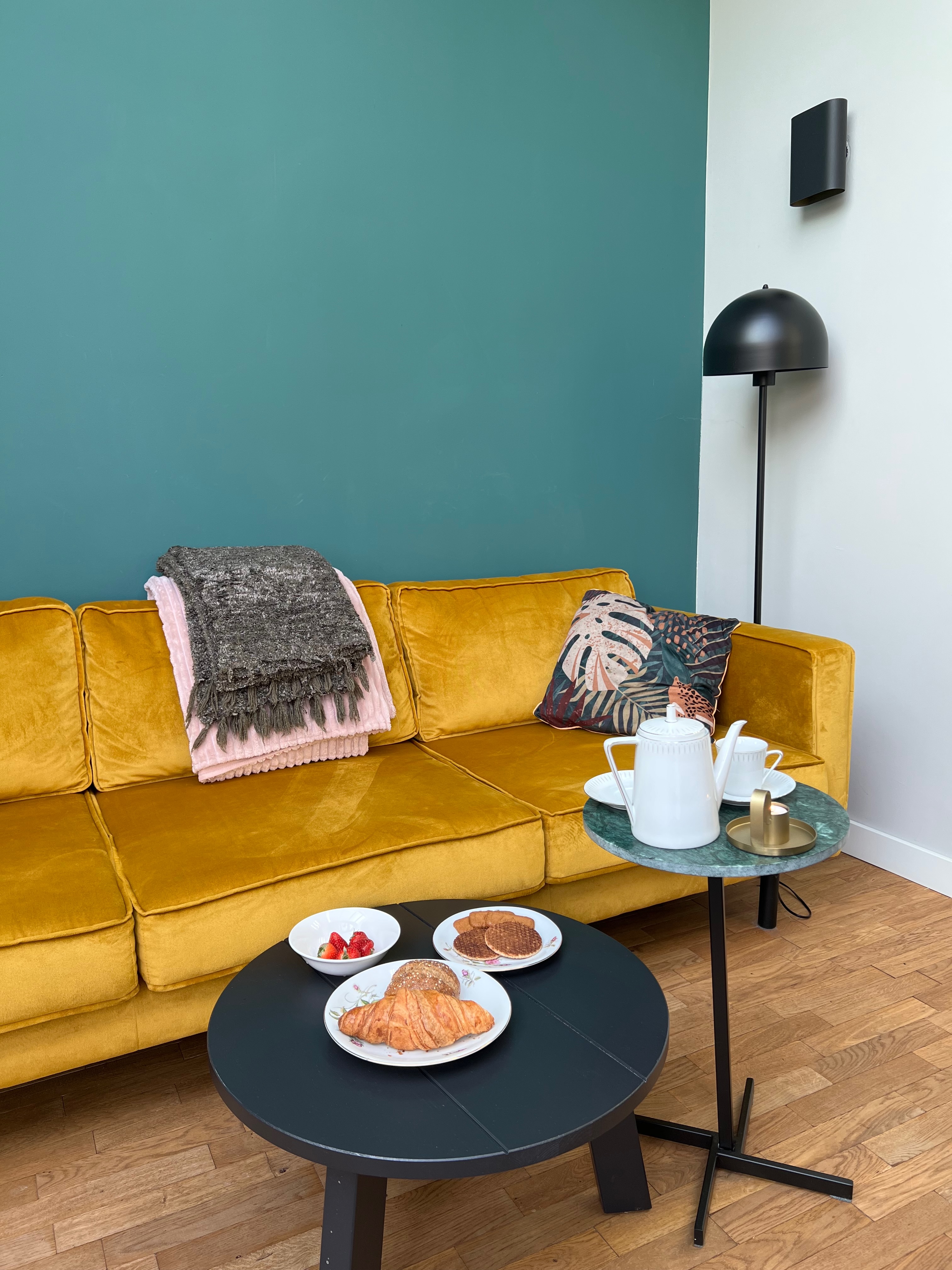 Breakfast spread from Airbnb: croissants, strawberries and stroop waffles lay on a table next to a teapot. The wall is a deep teal and the couch in the background is mustard-colored velvet.